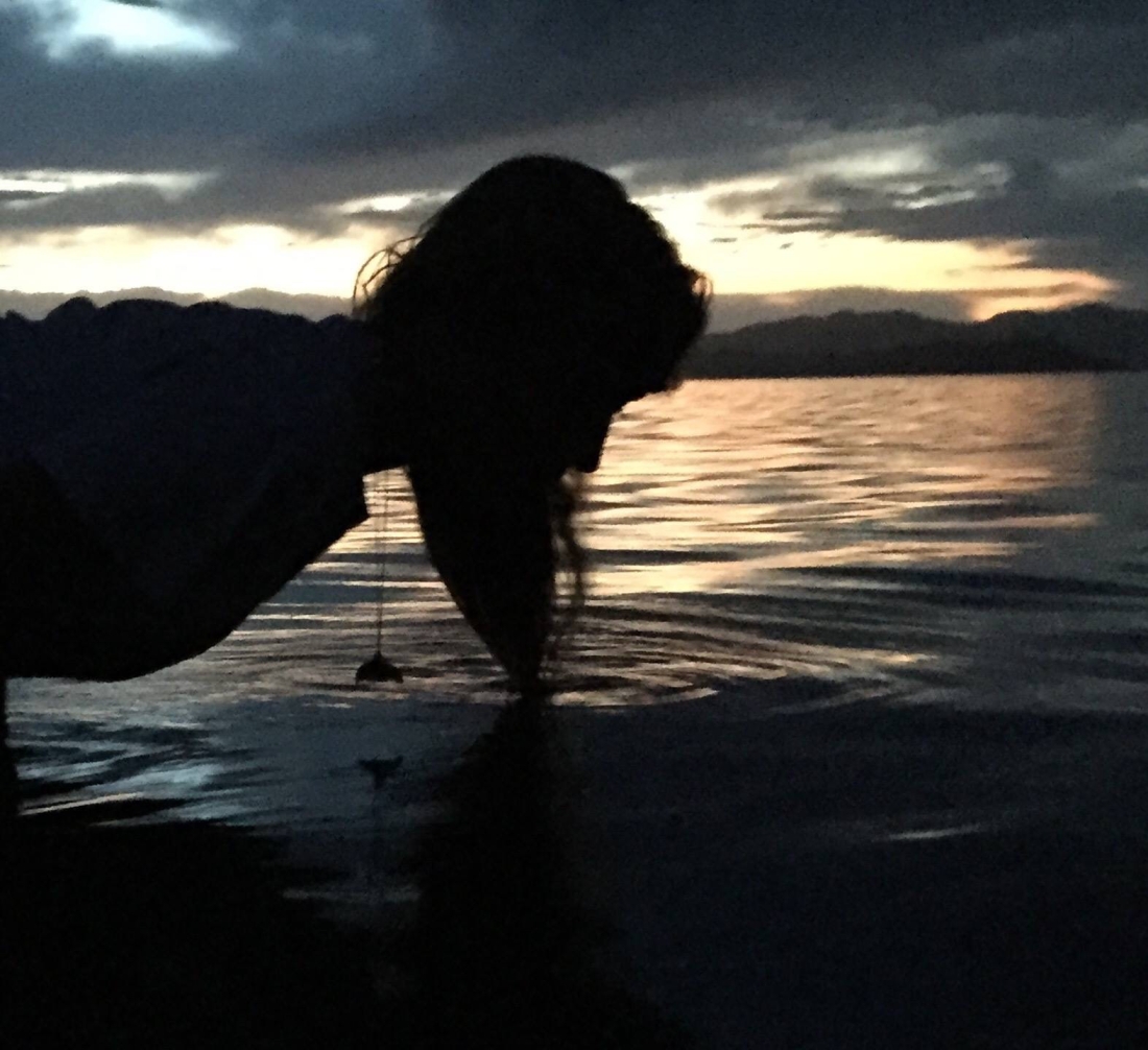 Silhouette of woman with the tip of her hair touching a lake. A sunset and dark clouds are behind her.